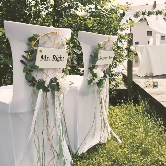Zwei dekorierte Stühle mit der Aufschrift Mr. und Mrs. Right stehen in der Wiese neben dem Holzsteg.